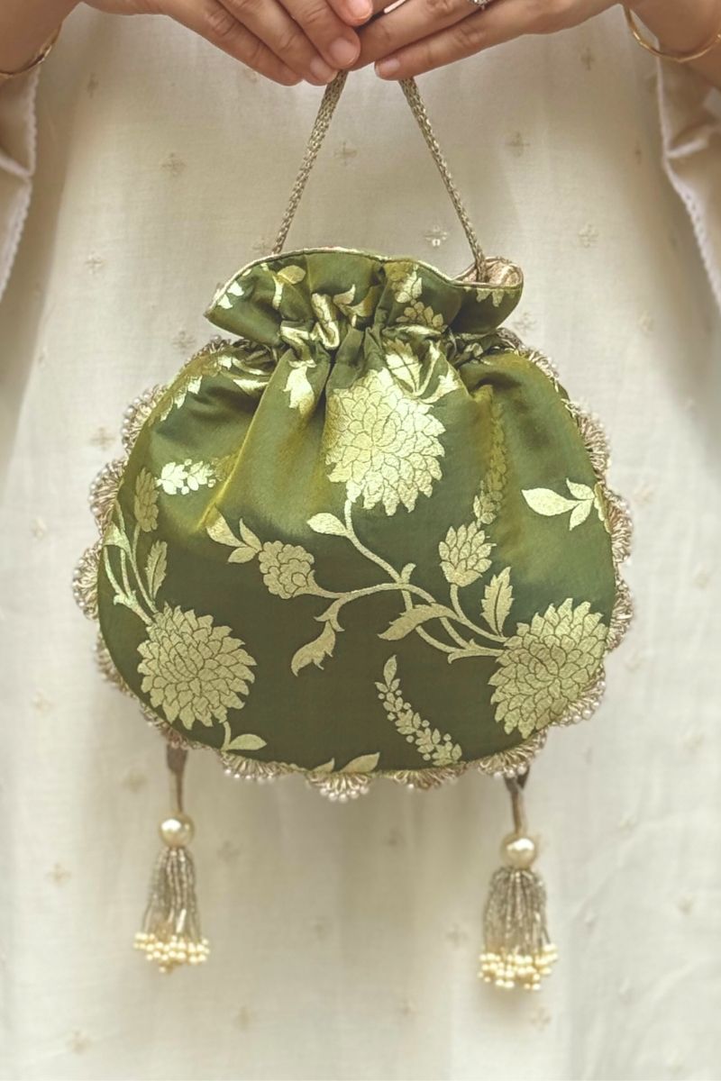 Hand holding elegant green Banarasi potli bag with gold floral motifs, scallop lace edges, and pearl tassels
