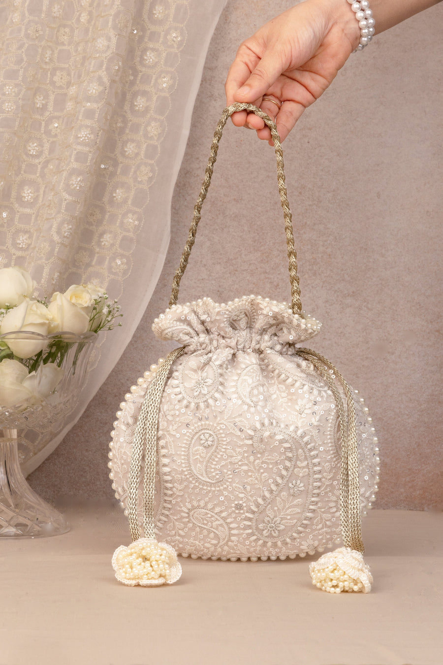 Cream potli bag with intricate embroidery and pearl tassels held by woven gold braided handle.