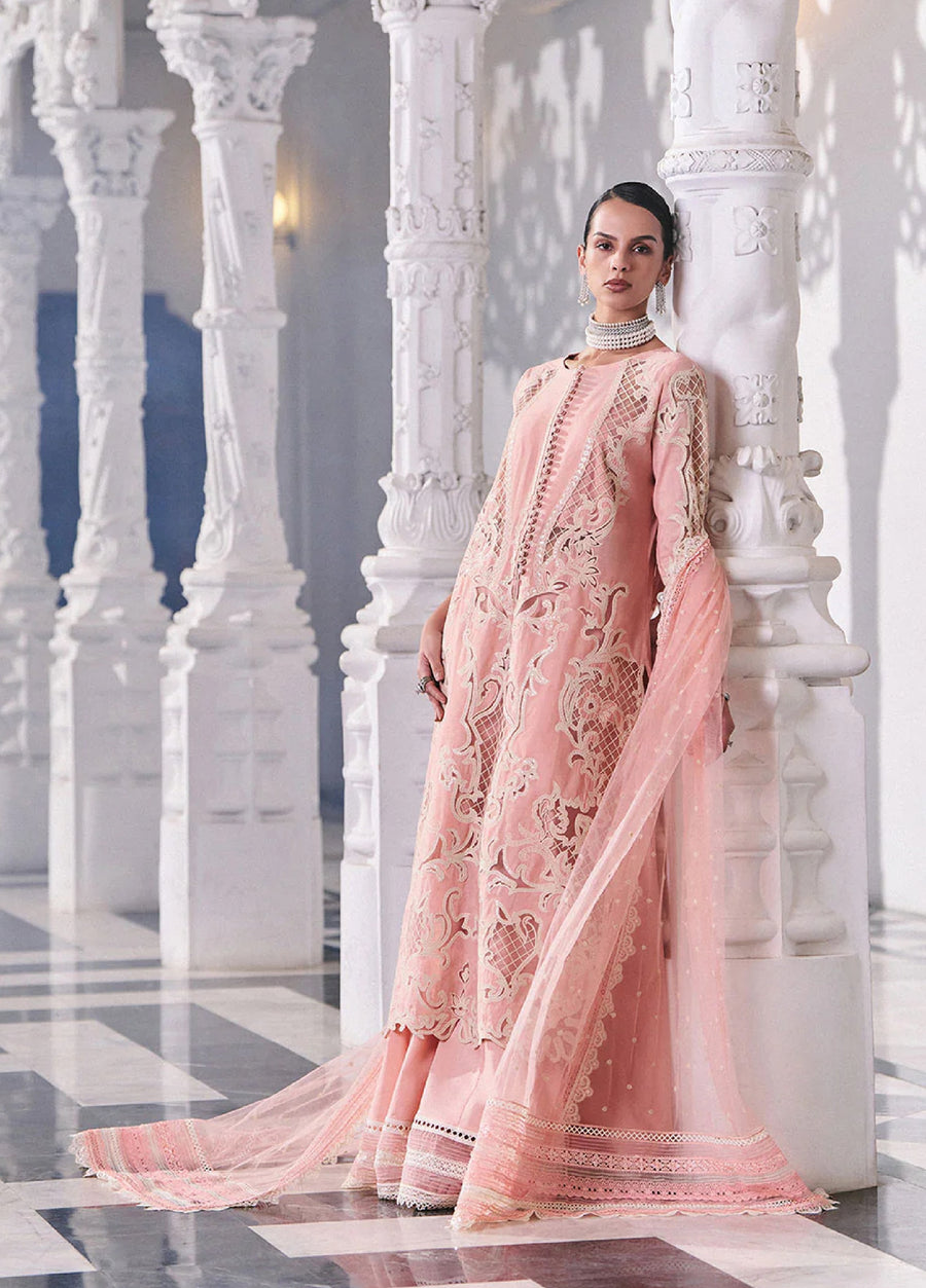 Model wearing an elegant soft pink embroidered lawn suit with intricate lace and embroidery, standing among white carved pillars in a luxurious hall.