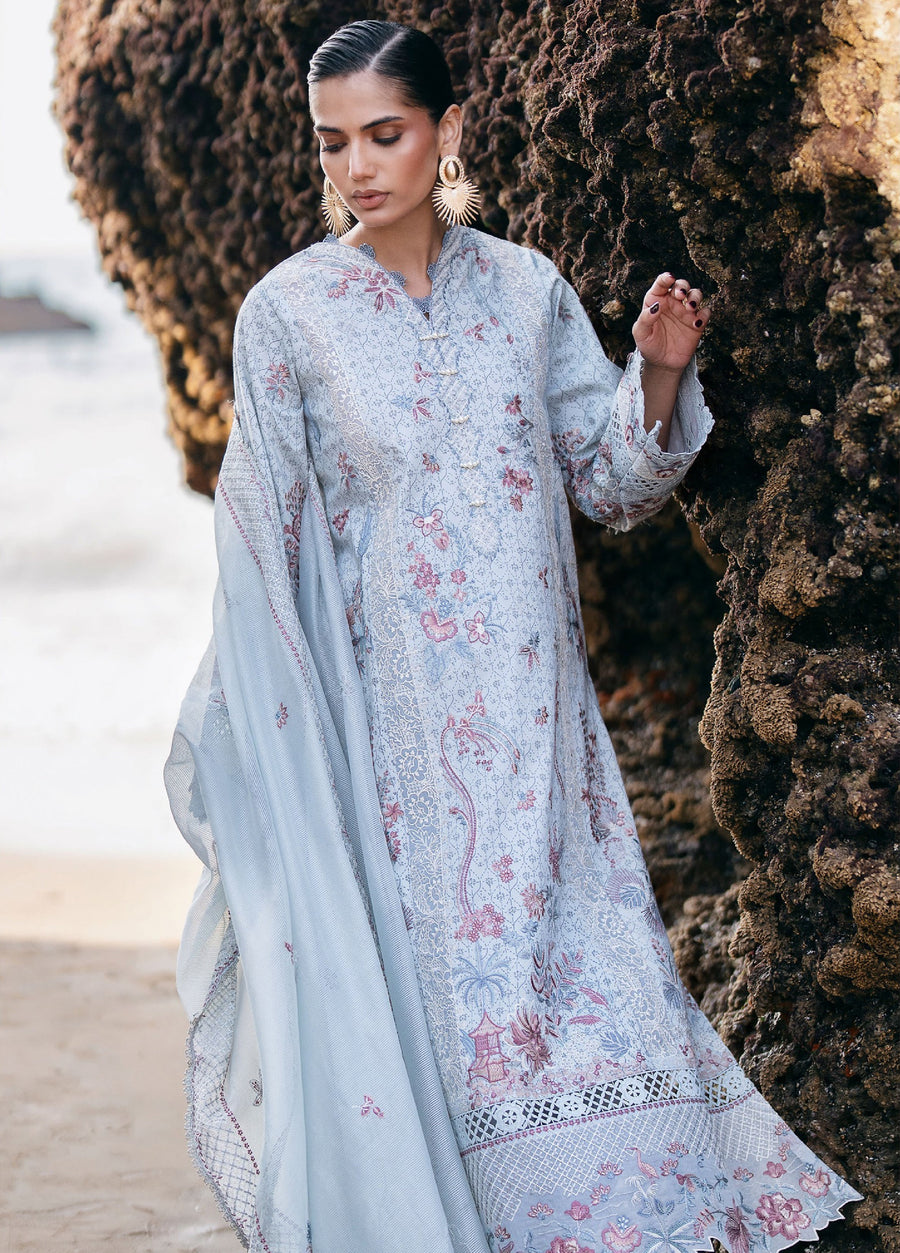 Woman wearing a pastel blue embroidered lawn suit standing near rocky seaside, showcasing floral embroidery and lace details on sleeves and hem