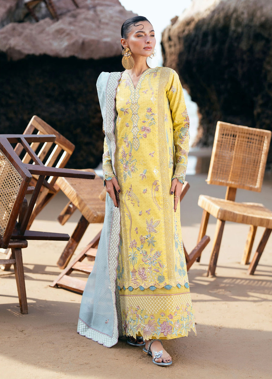Woman in a full-length yellow embroidered lawn suit with pastel floral motifs and a light blue embroidered dupatta draped over her shoulder, standing among wooden chairs on a sandy surface.
