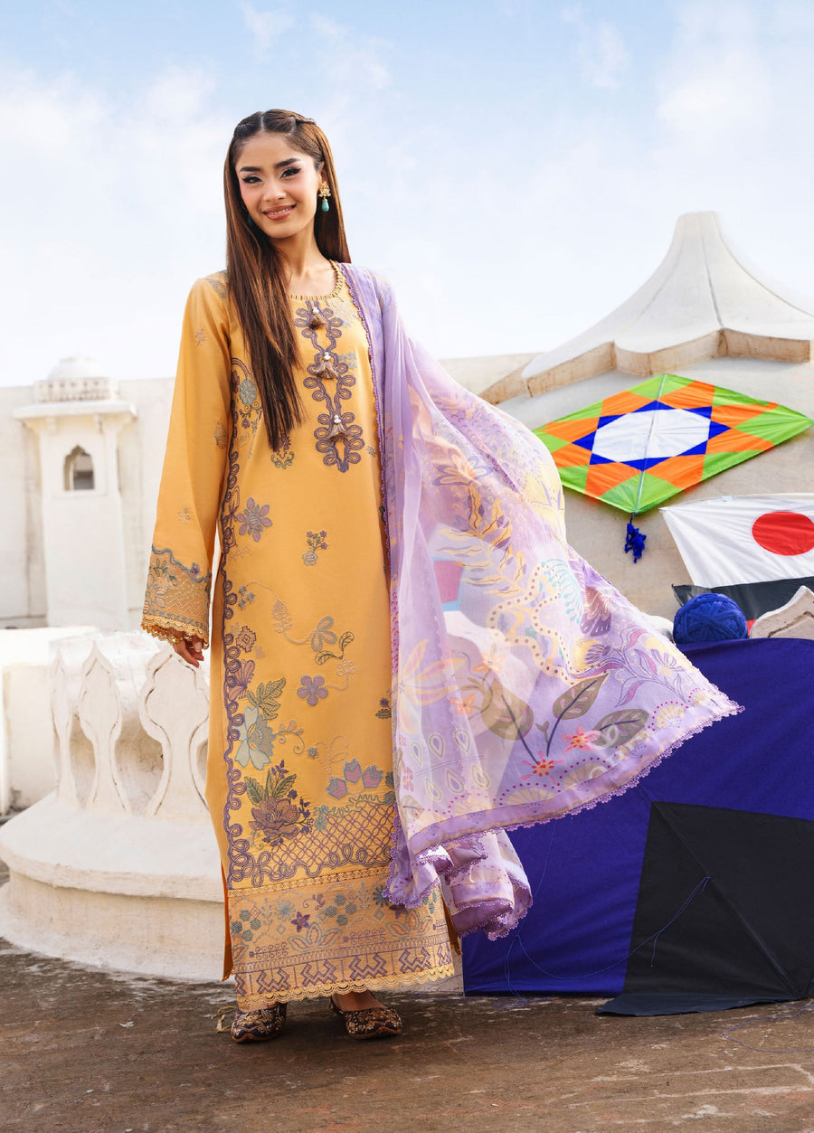 Woman wearing mustard yellow embroidered lawn shirt with lavender chiffon dupatta and patterned shoes, standing outdoors near white stone architecture and colorful kites