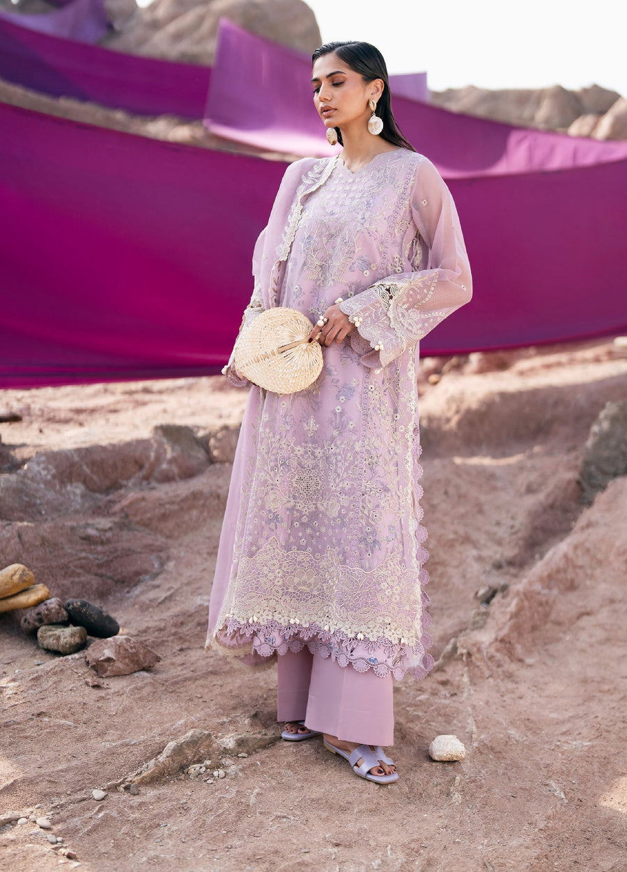 Woman wearing a soft lilac embroidered long shirt with intricate floral patterns and lace detailing holding a woven round handbag against a rocky background with purple fabric draped behind her.