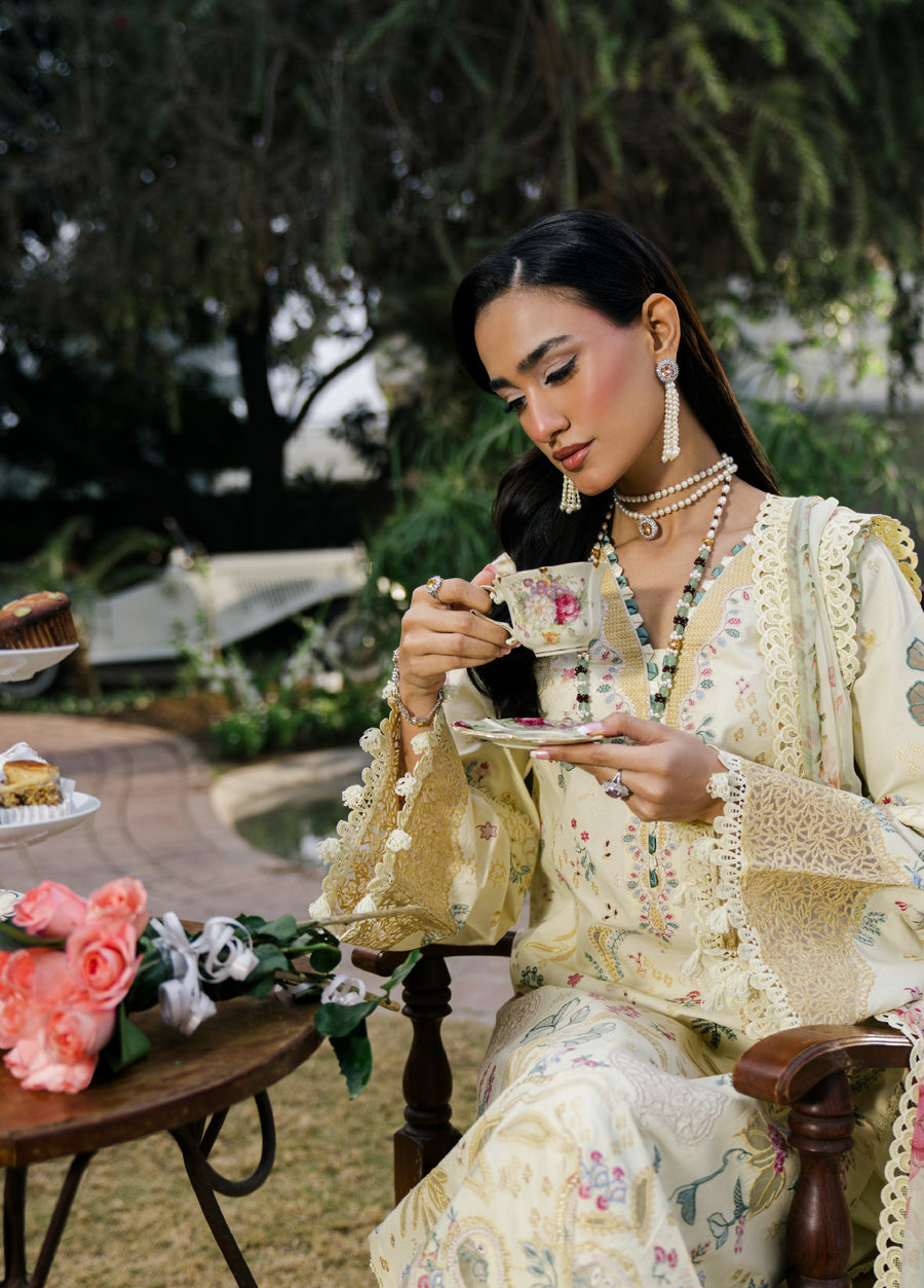 Close-up of model seated holding floral tea cup wearing embroidered suit with detailed lace cuffs and elegant jewelry