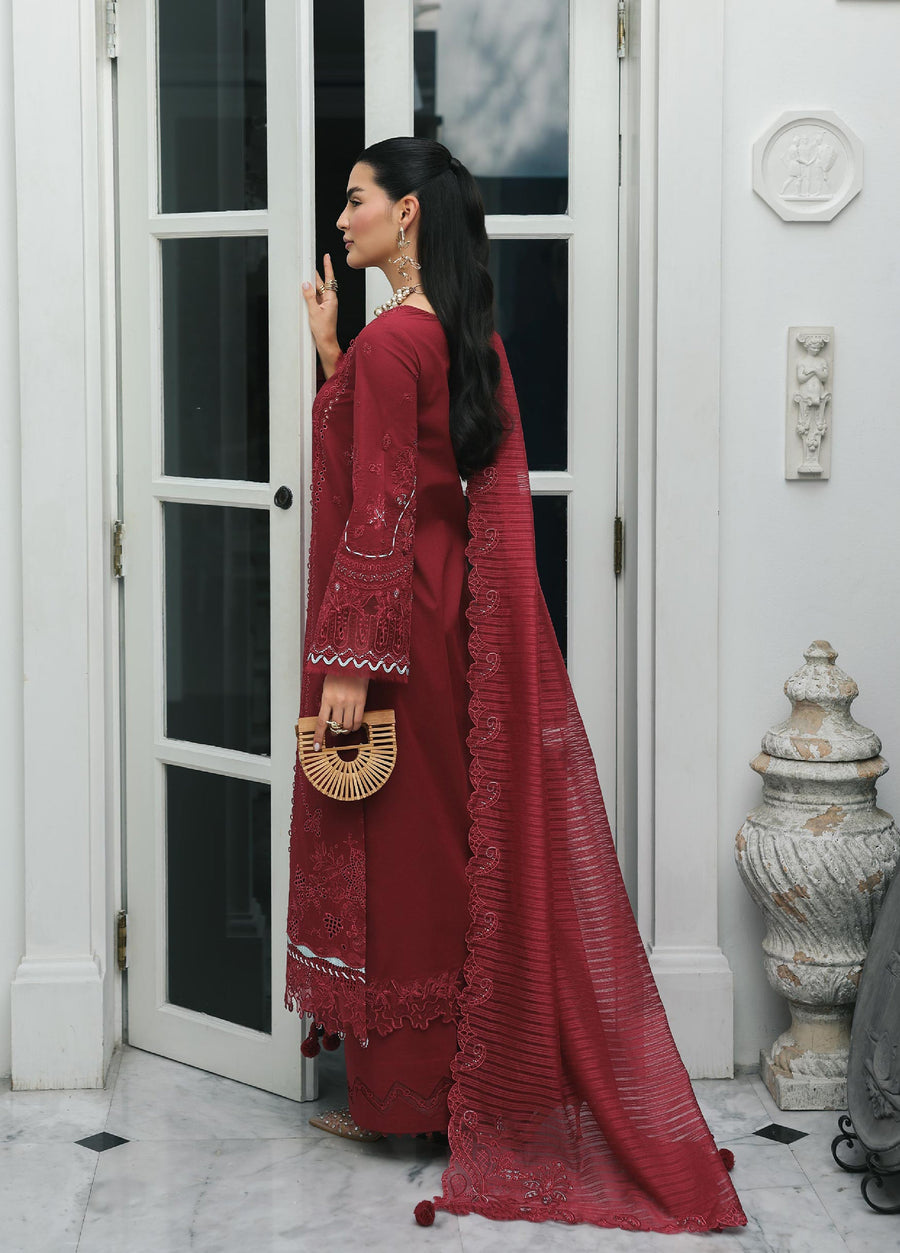 Side profile of a woman wearing a maroon embroidered lawn suit with matching trousers and a detailed dupatta, holding a wooden handbag, standing indoors near a door.