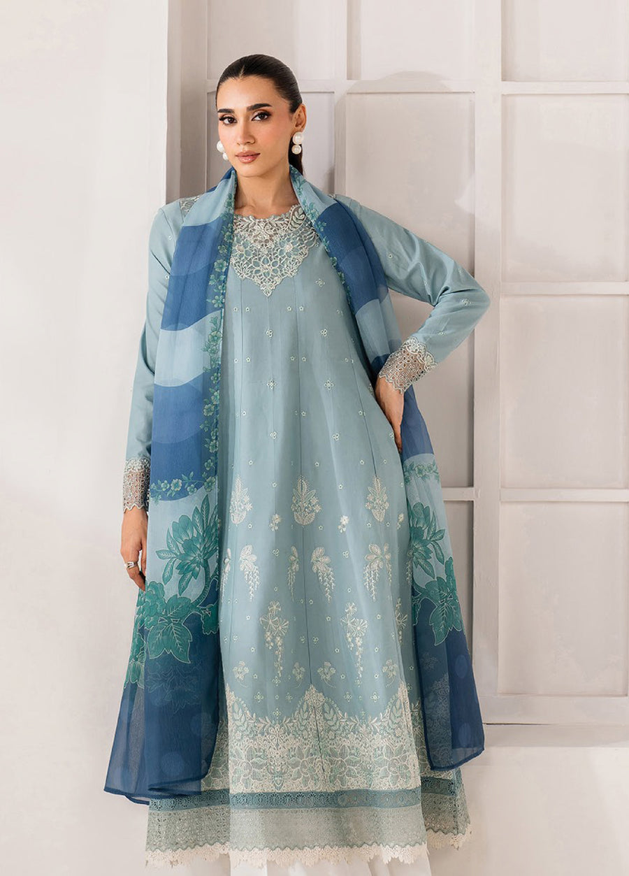 Woman wearing a sky blue embroidered lawn suit and matching blue printed dupatta, accessorized with pearl earrings, standing in front of a neutral background.