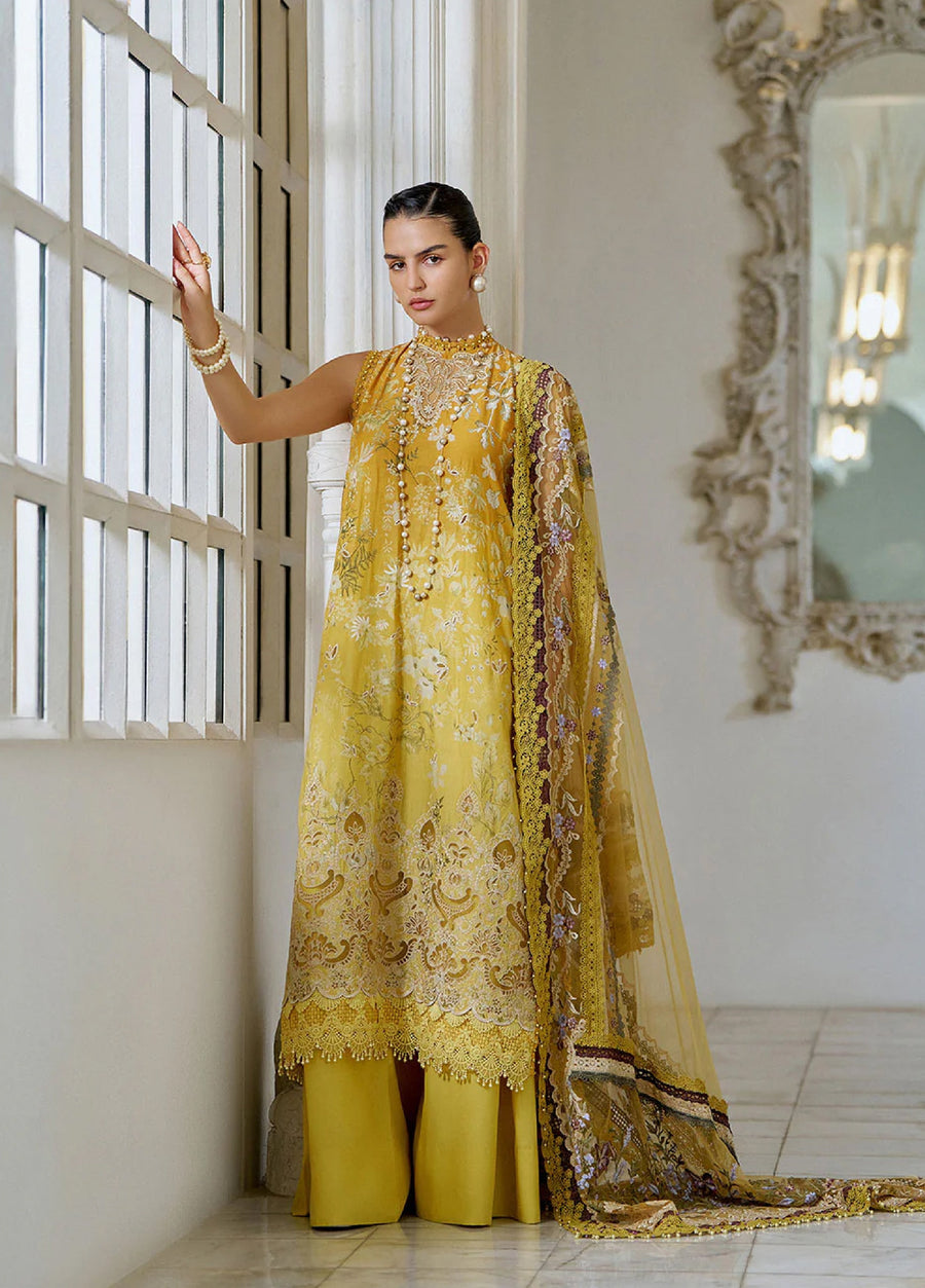 Three-quarter frontal view of the woman standing against a white wall wearing the mustard embroidered suit, highlighting the high neck, intricate floral embroidery, and pearl accessories.