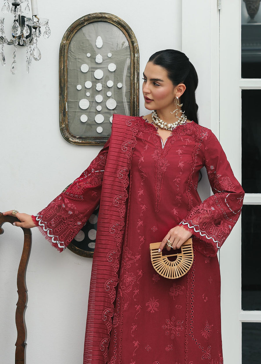 Close-up of a woman in a maroon embroidered lawn suit with intricate sleeve detailing and scalloped hems, accessorized with statement pearl jewelry and holding a wooden handbag indoors.