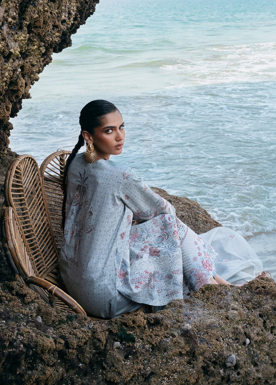 Woman seated by the seaside on rocks wearing pastel blue embroidered lawn suit, highlighting the back embroidery and soft net dupatta