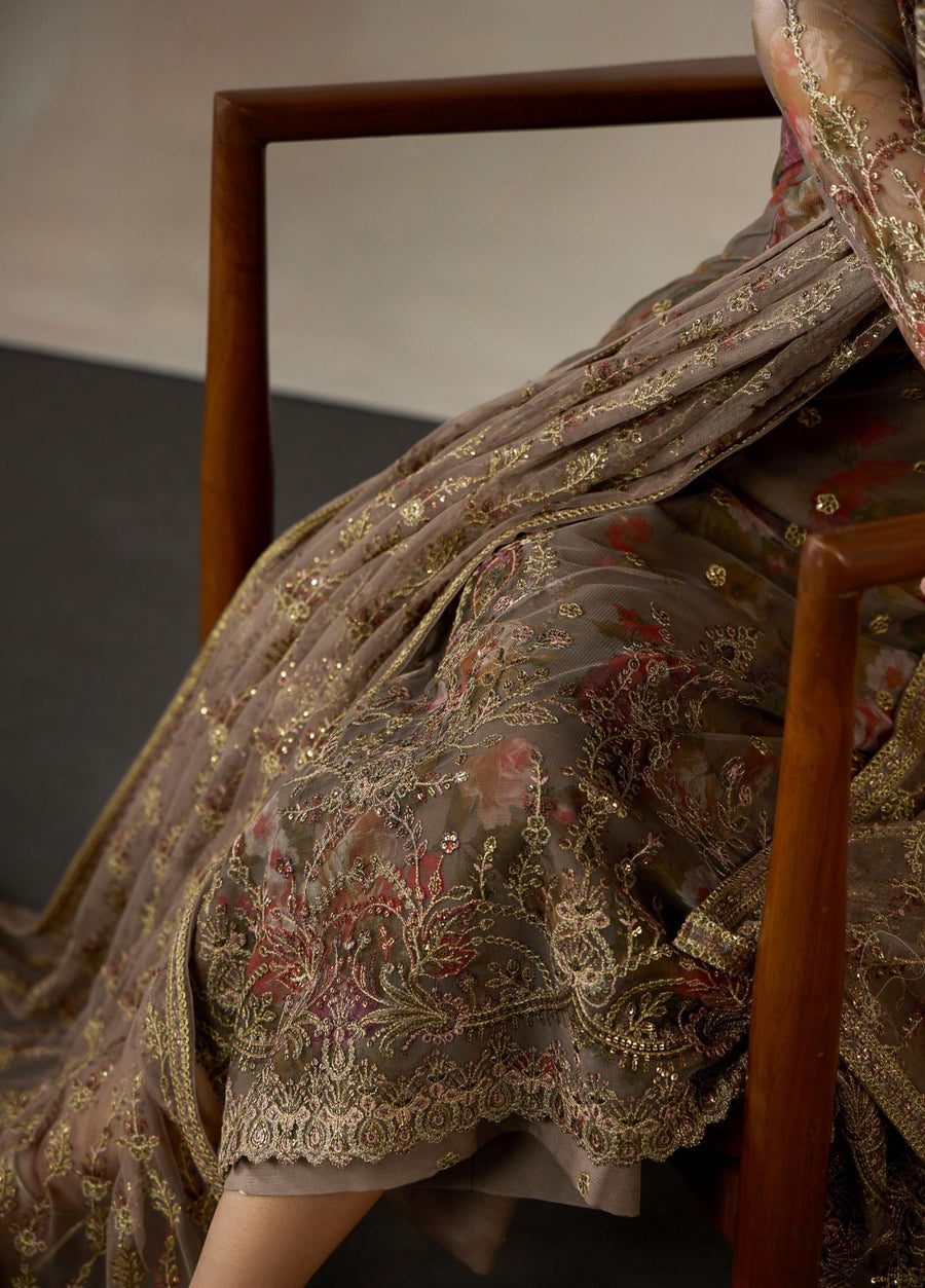 Woman seated beside wooden armchair adjusting golden earrings, wearing embroidered brown floral net suit with detailed dupatta
