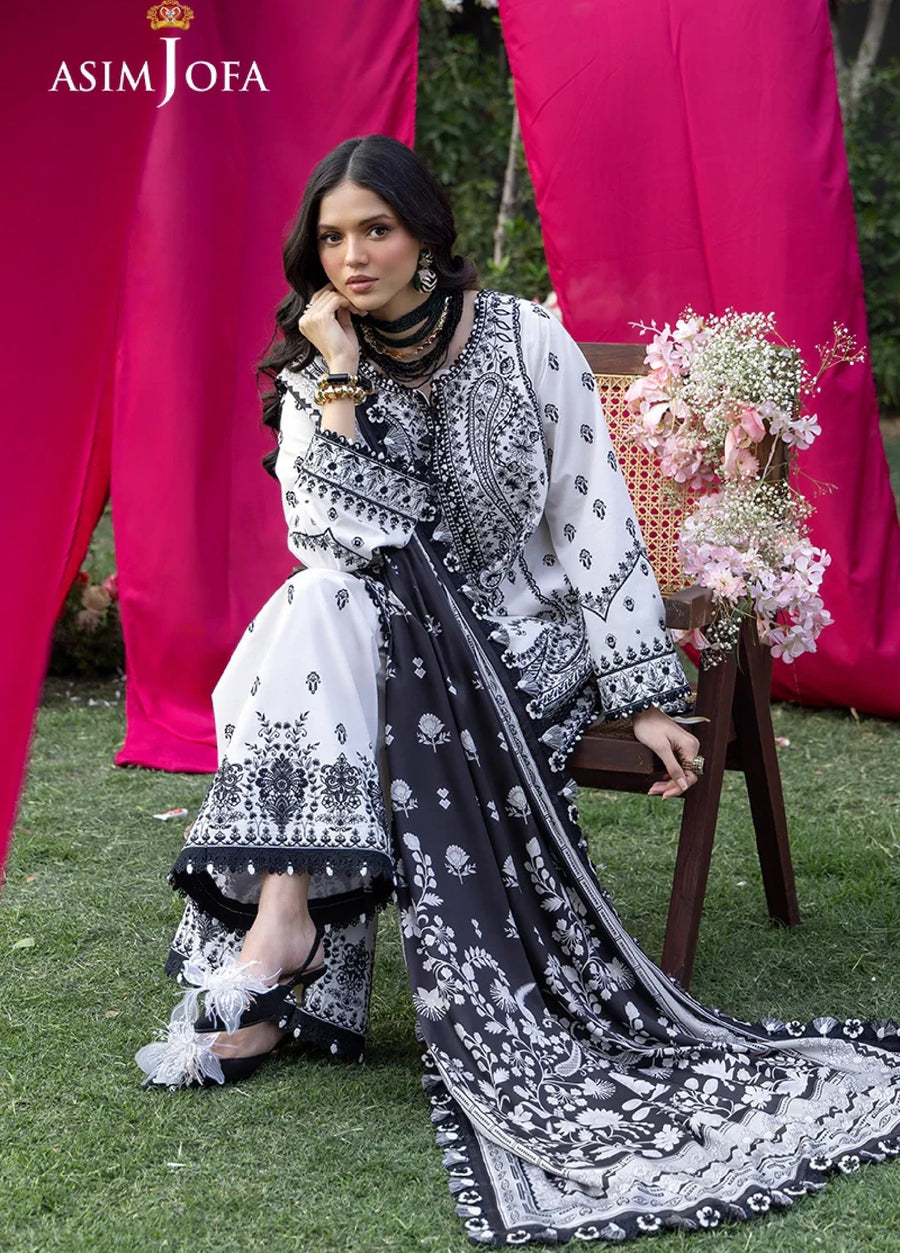 Seated woman dressed in black and white embroidered lawn suit with silk dupatta and black sandals, highlighted by floral and tassel accents.