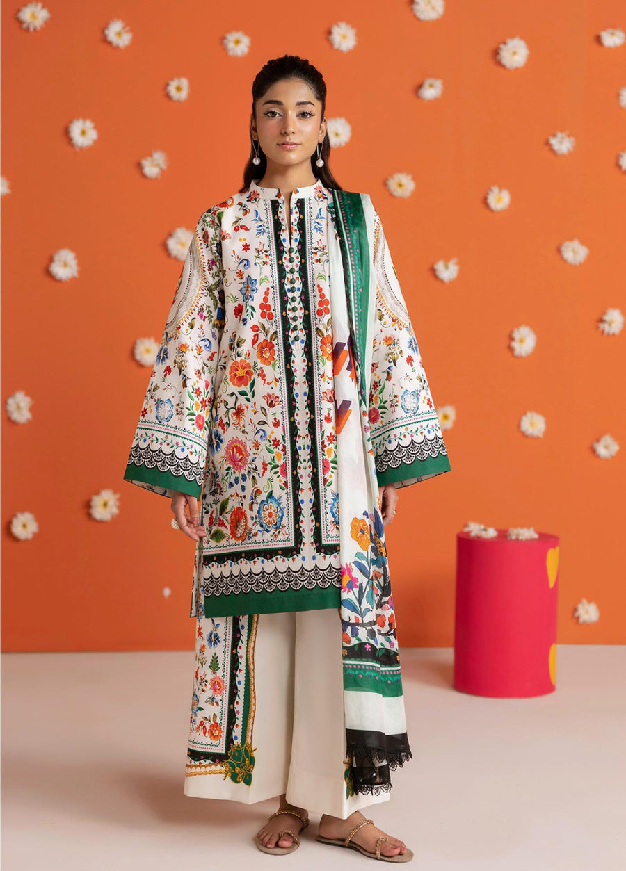 Front view of woman wearing a floral printed lawn 3-piece suit with green borders and vibrant multicolor floral patterns, standing against an orange wall decorated with white flowers.
