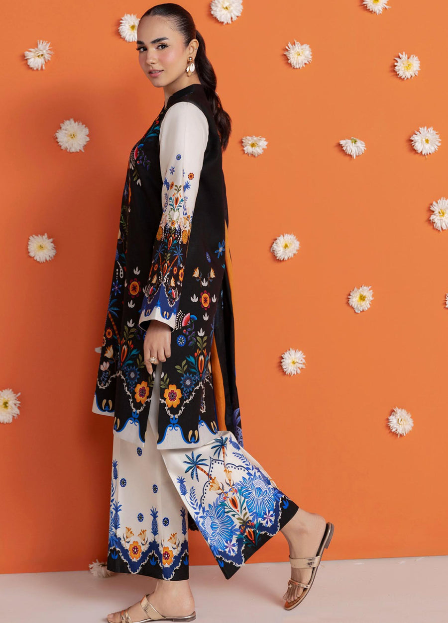 Side view of a woman modeling a black lawn suit with detailed floral prints, cream trousers with blue motifs, and a matching dupatta on an orange floral backdrop