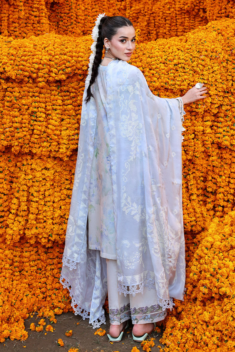 Rear view of woman wearing Lilac Whisper suit showing multi-patterned silk dupatta and floral embroidered borders on trousers against marigold flower wall