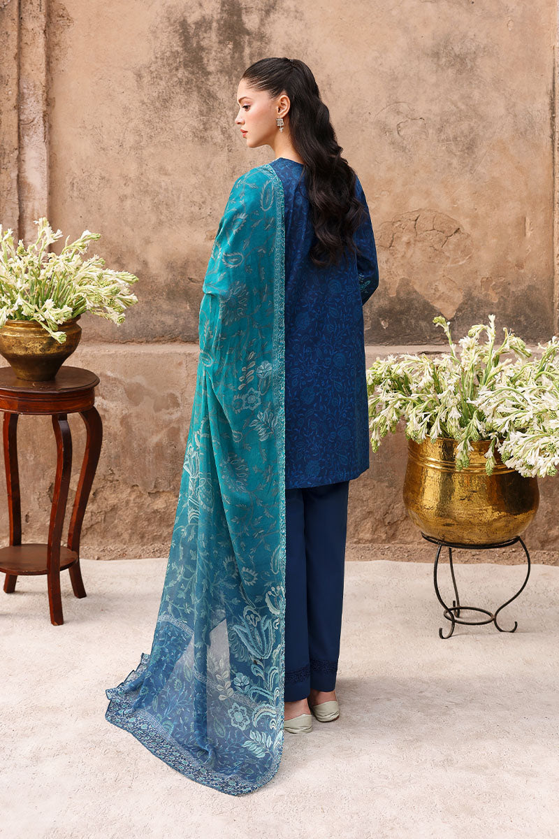 Back view of woman in navy blue embroidered lawn suit with teal dupatta against a rustic wall background