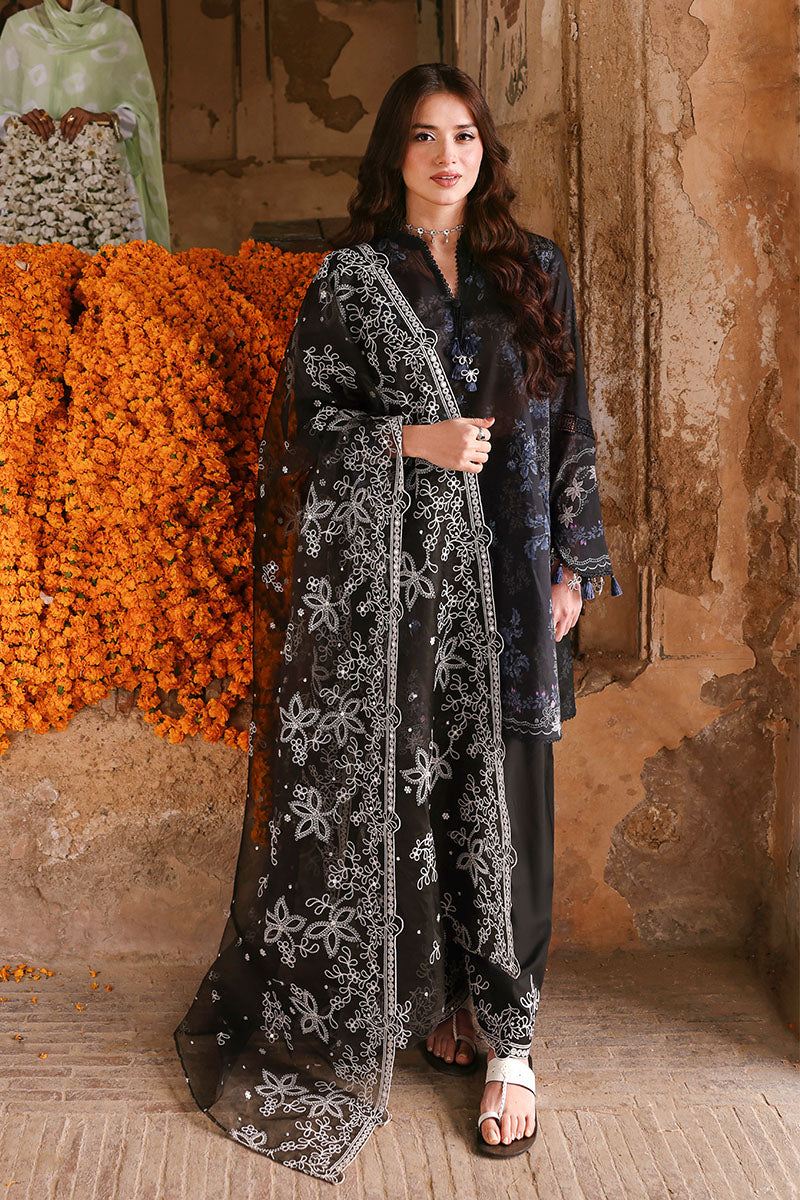 Woman standing confidently in black embroidered lawn suit with detailed white embroidered organza dupatta draped over shoulder, orange marigold flowers behind her.