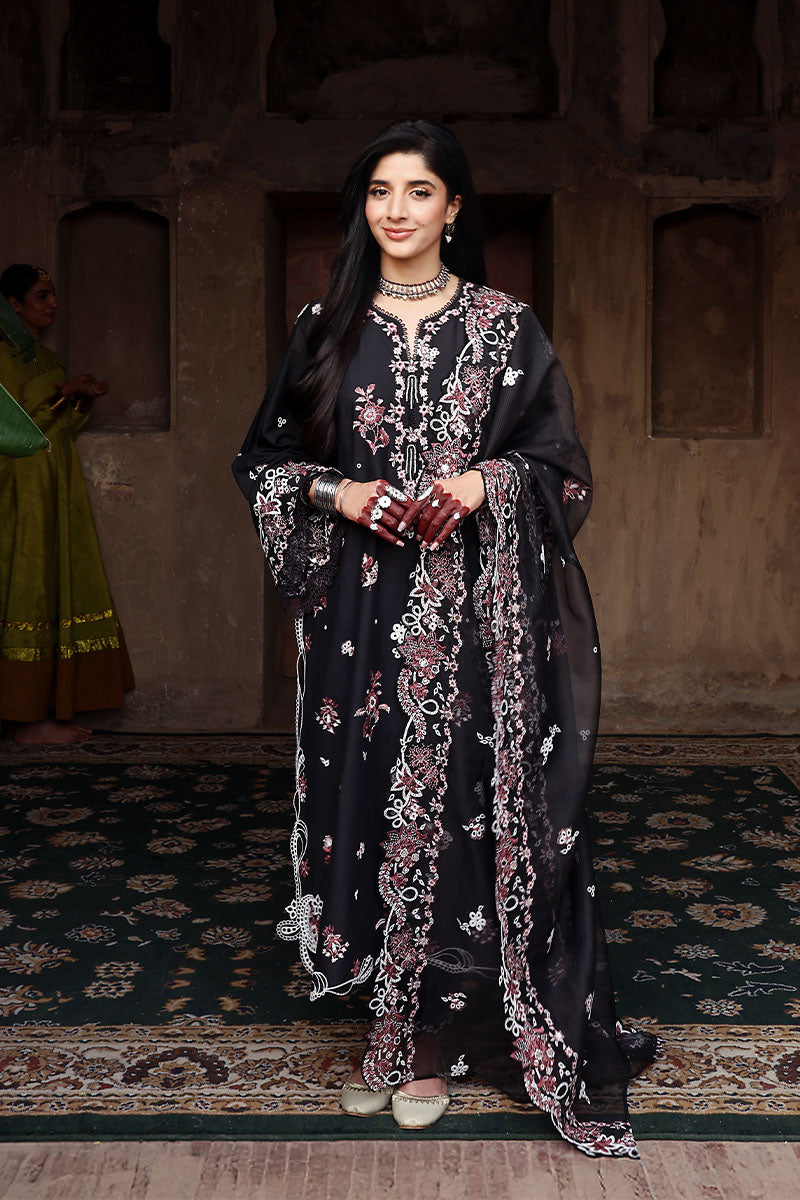 Woman wearing a full black lawn suit with intricate rose and white floral embroidery, posing indoors on a patterned carpet.