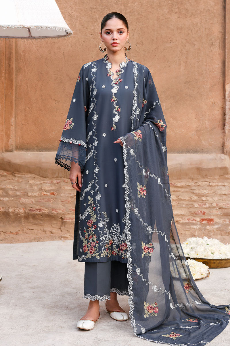 Woman wearing a dark slate blue embroidered formal lawn suit with floral embroidery, holding a matching sheer embroidered dupatta, standing against a rustic brown wall