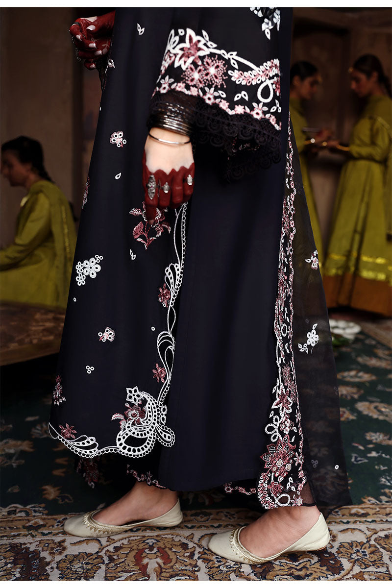 Close-up of the woman’s hand adorned with rings and bangles with embroidered black lawn suit and dupatta featuring floral and lace details, visible against traditional floor décor.