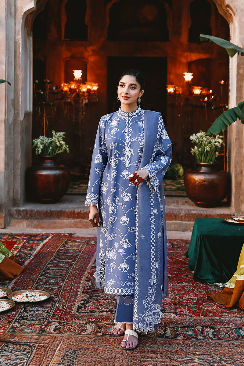 Full view of woman standing in blue embroidered lawn suit featuring floral patterns and matching net dupatta, set against a heritage backdrop