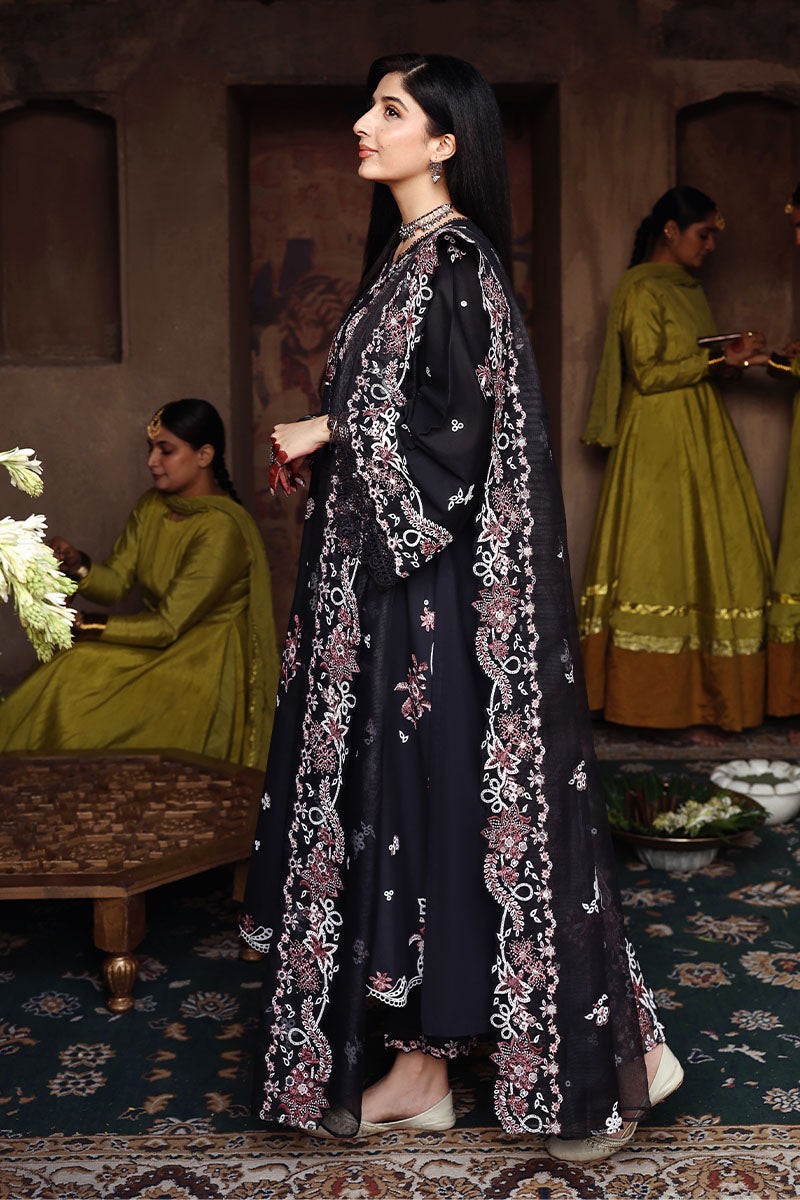 Side profile of woman in black embroidered lawn suit with full sleeves and matching dupatta, set indoors with background figures wearing green outfits.
