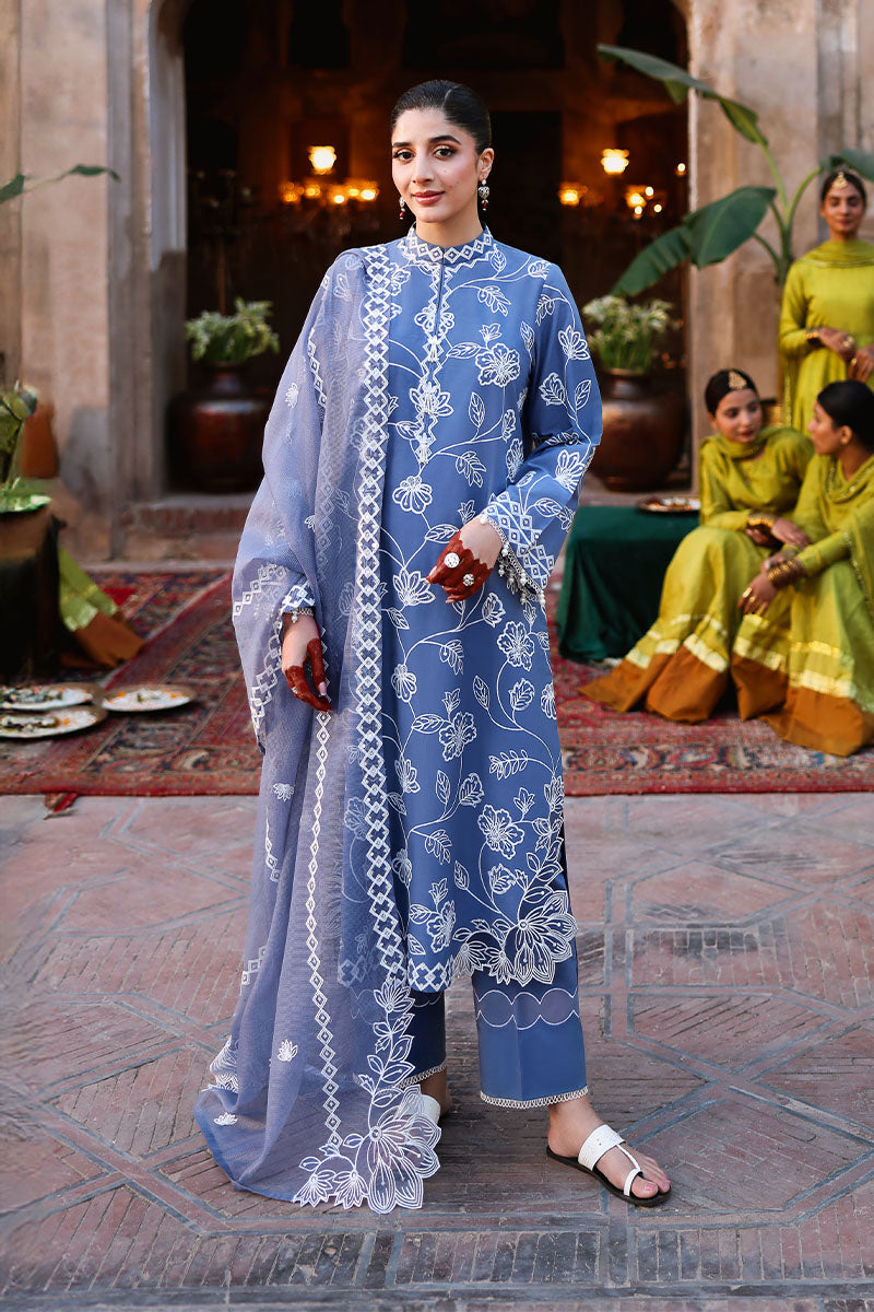 Woman wearing a blue embroidered lawn suit with floral and geometric designs, styled with a net dupatta, standing outdoors near traditional décor