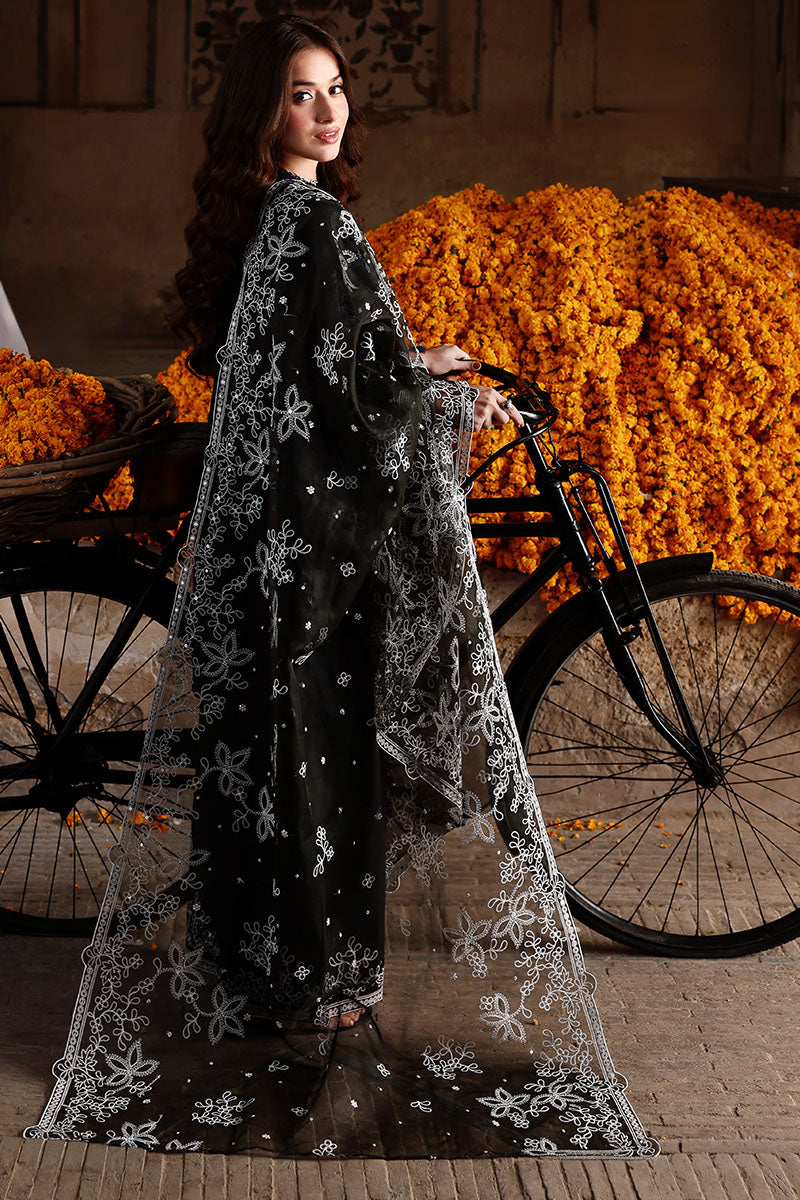 Back view of woman in black embroidered lawn suit holding sheer organza dupatta, with a bicycle and vibrant orange marigold flowers in the background.