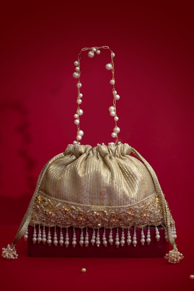 Gold potli bag with intricate floral embroidery, pearl tassel fringes, and pearl-beaded gold chain handle on red background