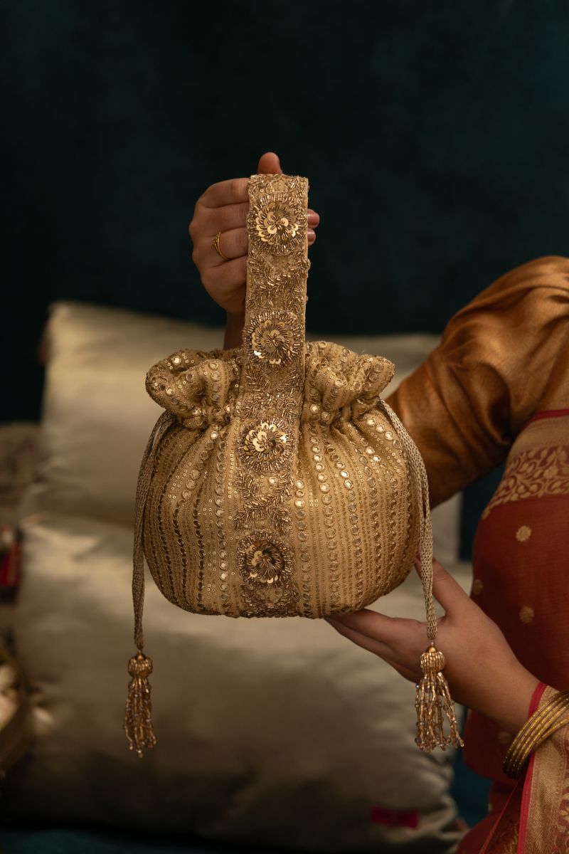 Hand holding elegant round gold potli bag with intricate embroidery and sequins detailing, featuring a wide floral embroidered handle and decorative tassels.