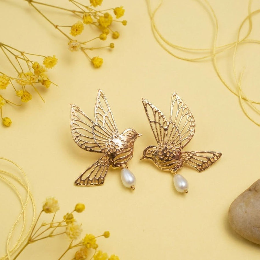 Gold bird-shaped earrings placed on a soft yellow background with small yellow flowers and light strings