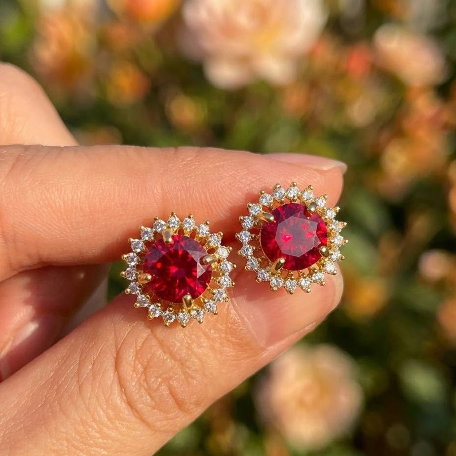 Hand holding red gemstone halo stud earrings with blurred flower garden background