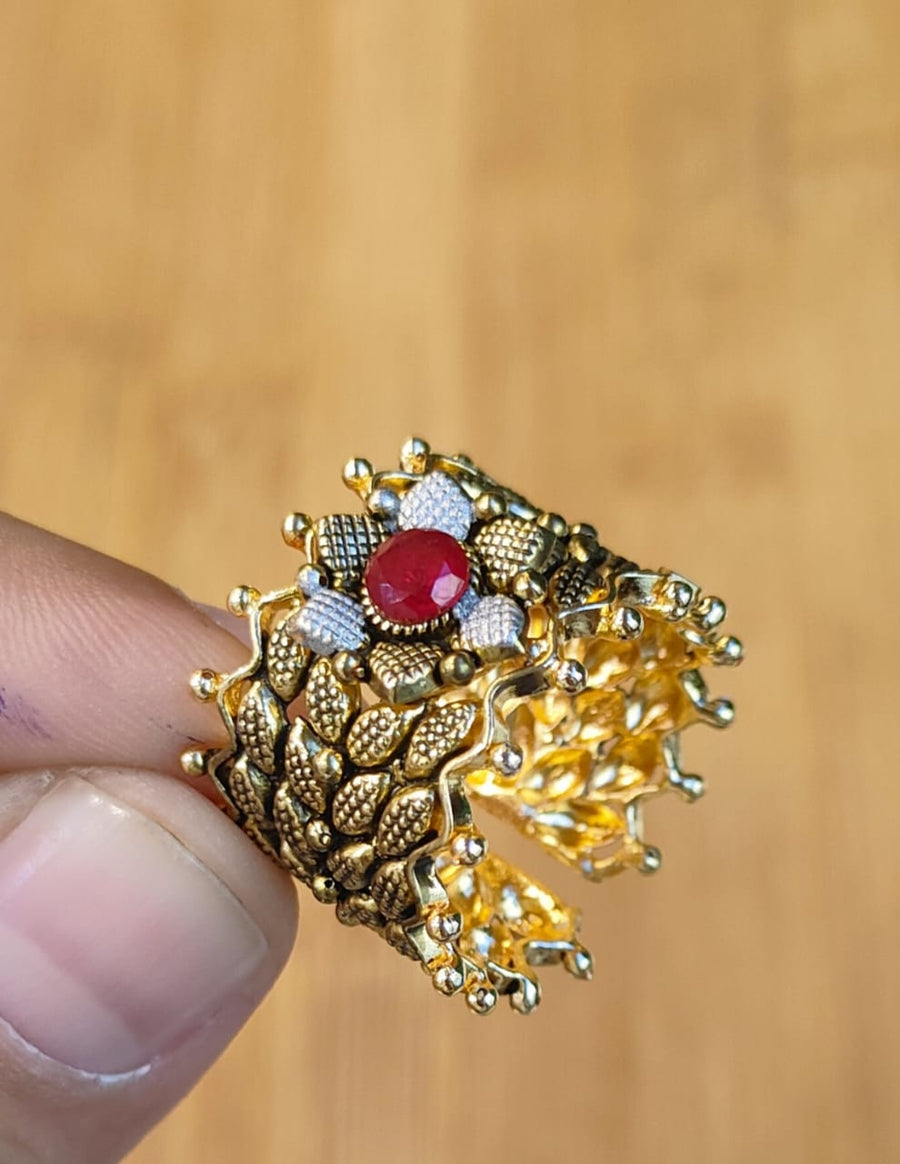 Detailed view of an adjustable wide gold-tone ring with textured design and red gemstone, held between thumb and forefinger against a wooden background.