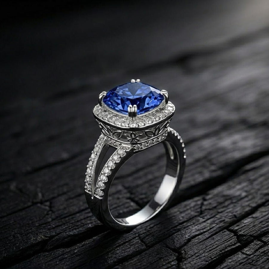 Close-up of cushion-cut blue gemstone ring with double pavé halo and floral engravings on dark textured surface