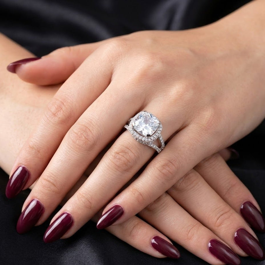 Women's hand wearing cushion-cut clear gemstone pavé ring with dark red manicure
