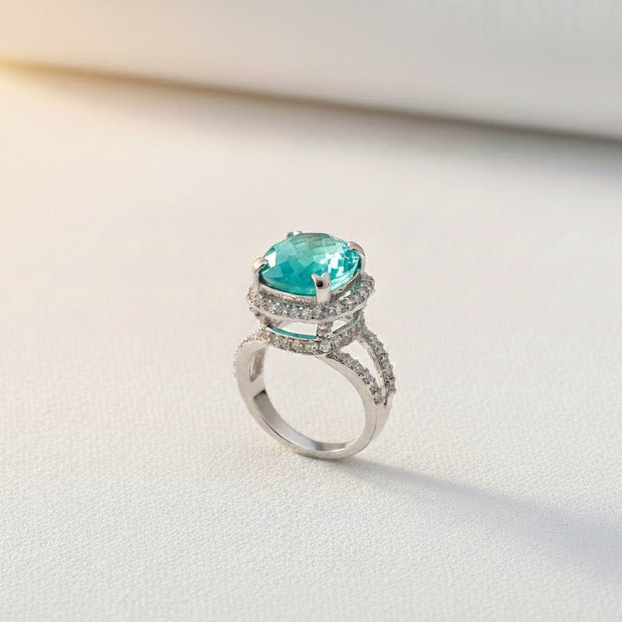 Teal cushion-cut gemstone ring with double pavé halo and split silver-tone band on light background