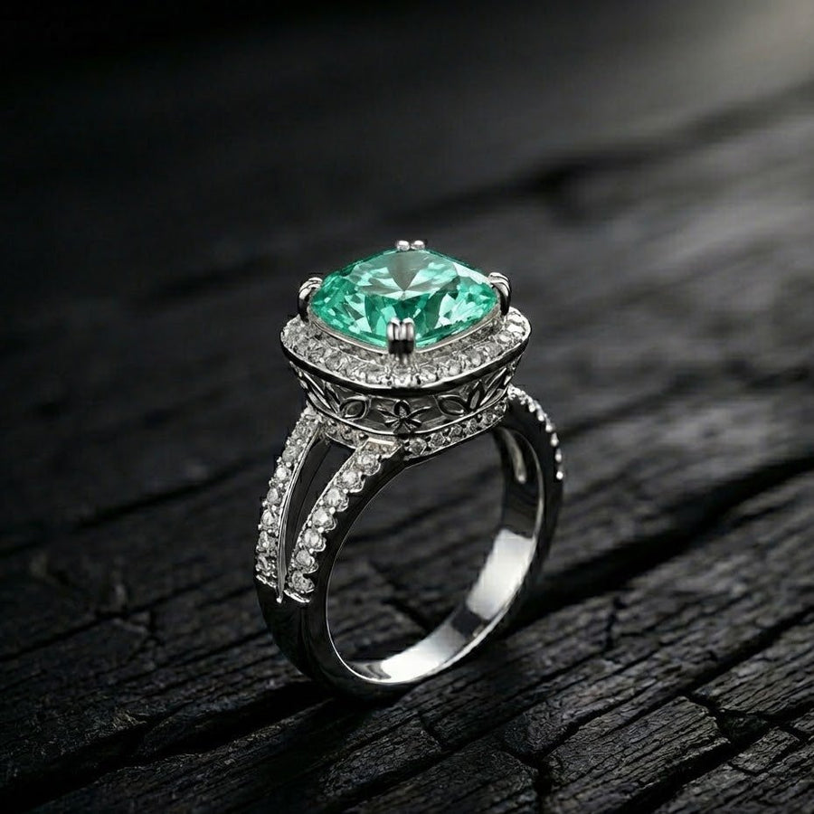 Close-up of cushion-cut green gemstone ring with double pavé halo and floral engravings on dark textured surface