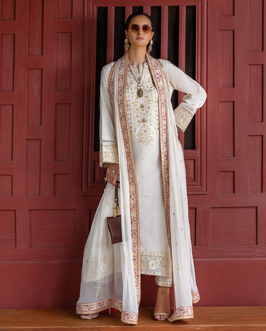 Woman wearing an embroidered ivory raw silk suit with gold and red detailing, standing in front of a red wooden door.