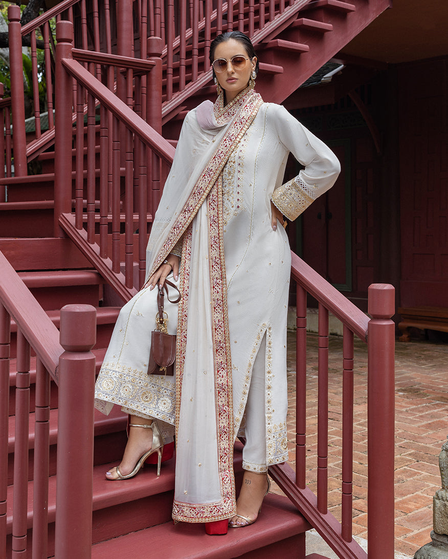 Side view of woman posing on red wooden stairs wearing embroidered raw silk suit with matching dupatta and high heels.