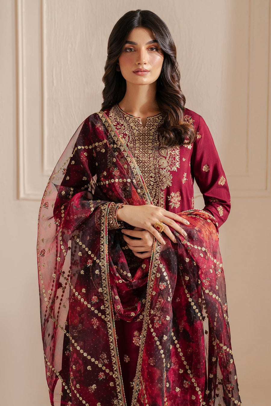 Portrait-style image of a woman wearing a maroon embroidered formal suit with delicate gold embroidery, styled modestly with a sheer organza dupatta draped elegantly across her arms.