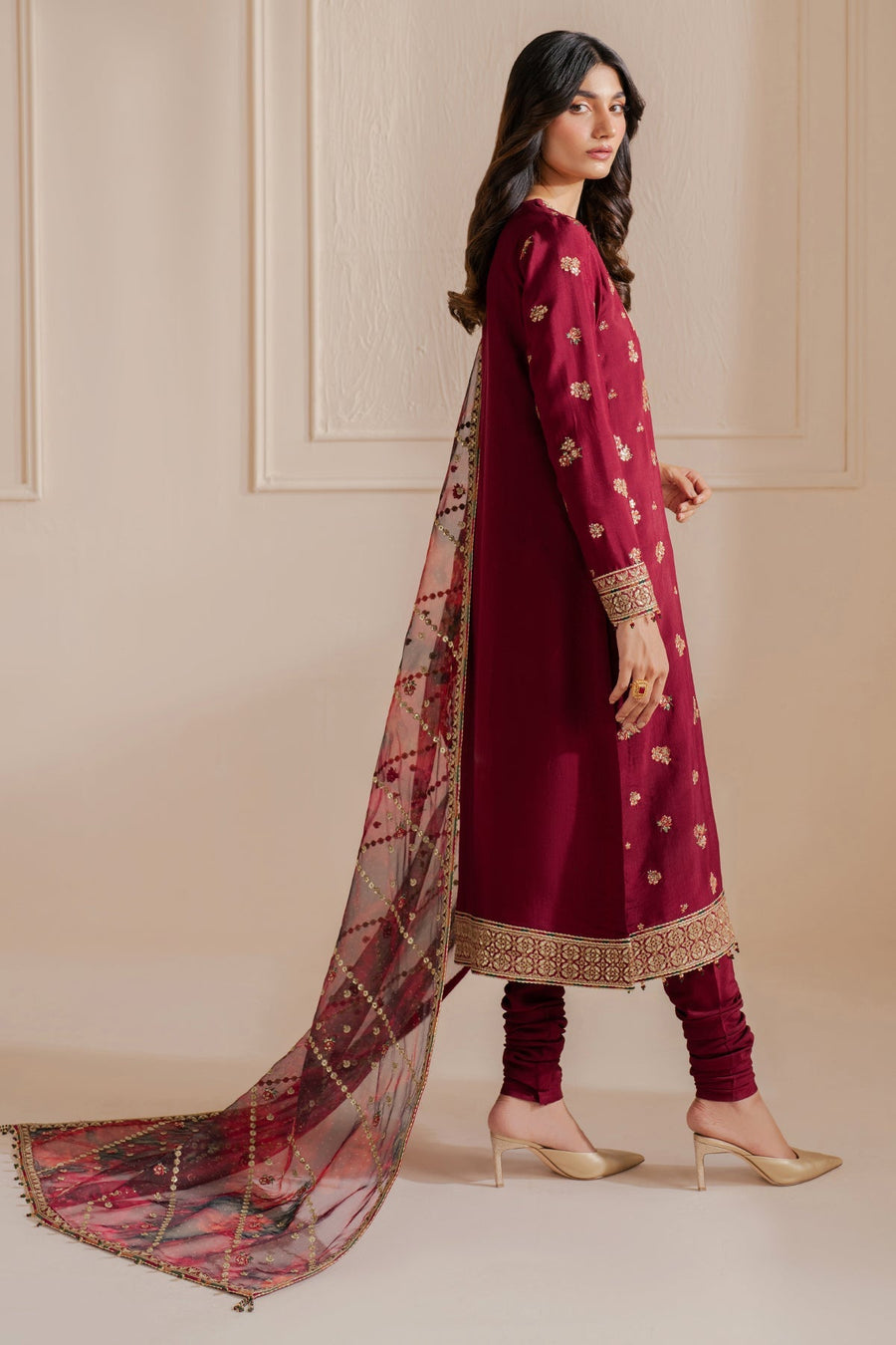 Side view of a woman dressed in a maroon embroidered suit featuring intricate gold embroidery on sleeves and hem, showcasing the long sheer organza dupatta trailing behind her, paired with beige high heel shoes.