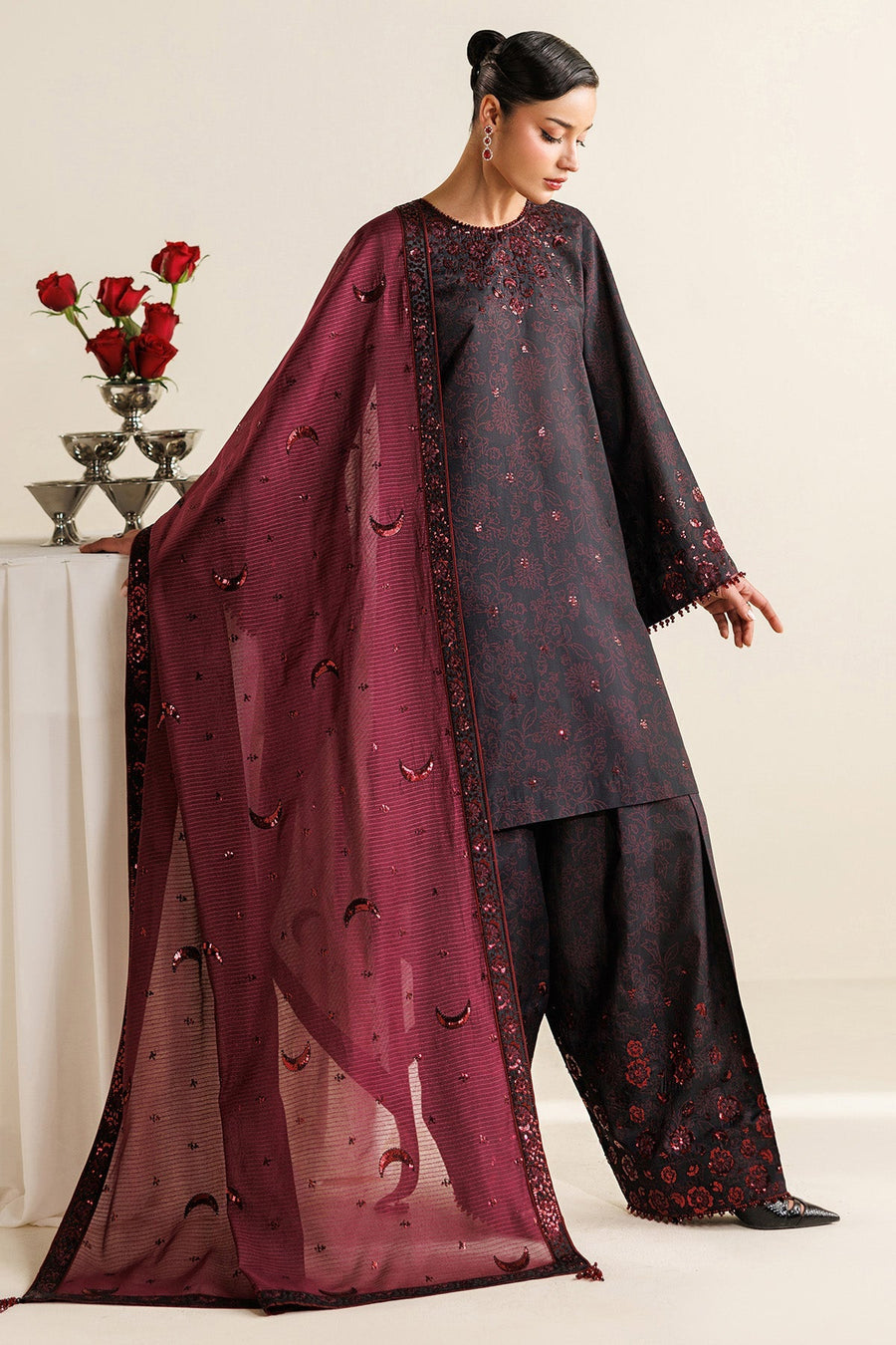 Model wearing elegant embroidered black kameez with maroon floral designs and matching wide-legged trousers, holding maroon dupatta with crescent motifs.