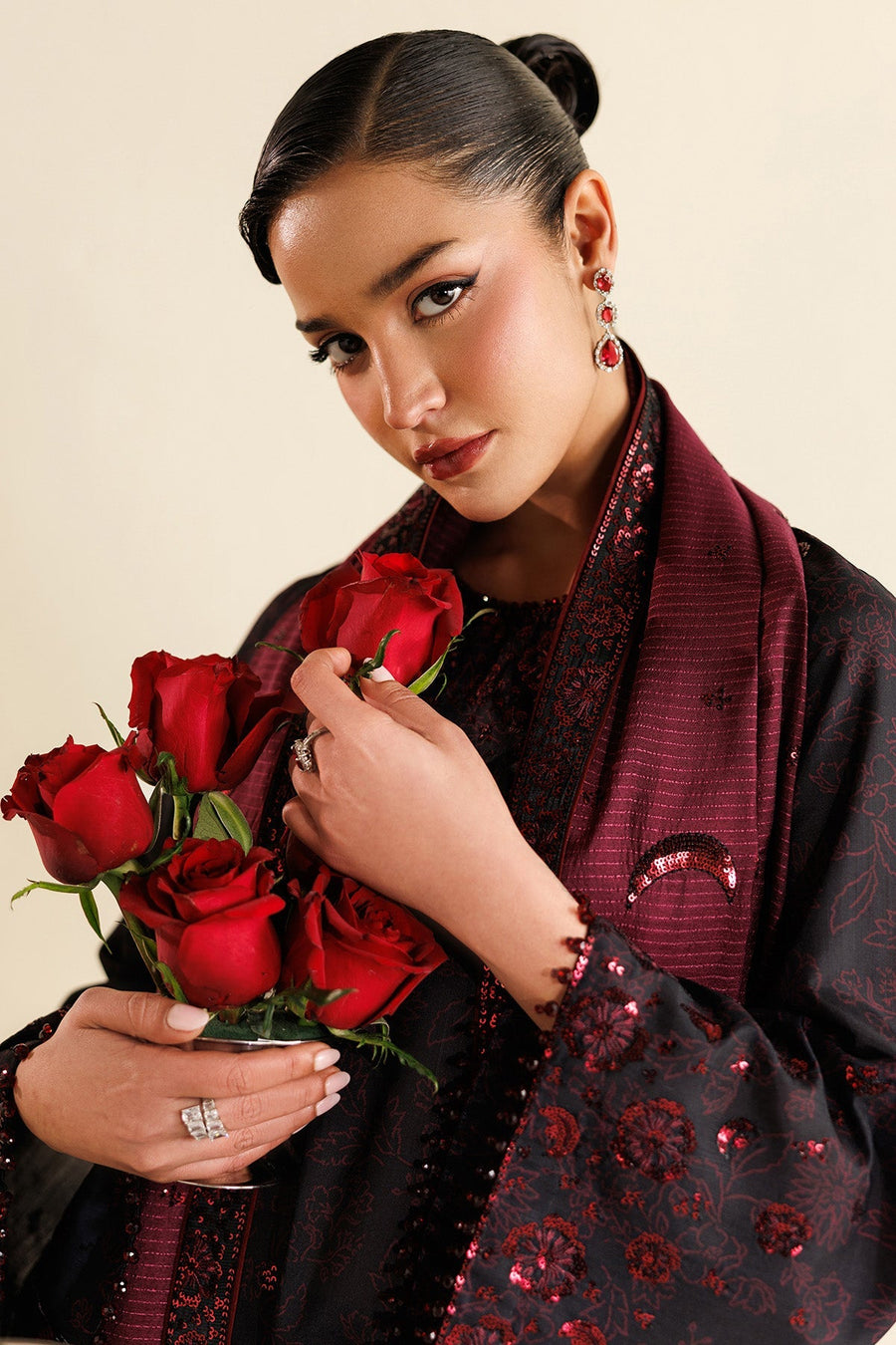 Model with maroon dupatta wrapped around shoulders, standing near decorative silver bowls, wearing embroidered black suit with floral sequin details.