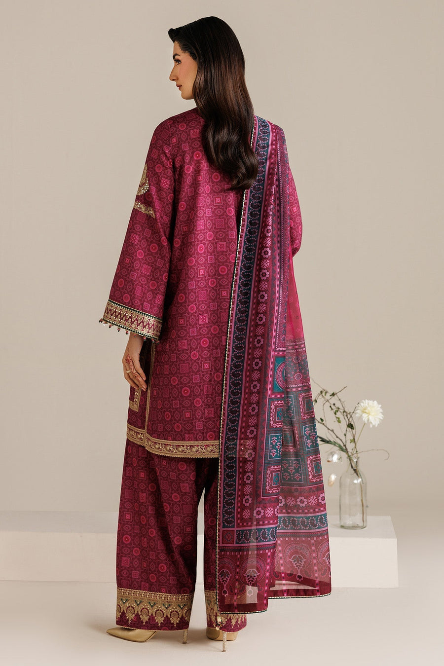 Model sitting gracefully wearing embroidered deep magenta suit and printed dupatta, showcasing embroidery on trousers and border details, accessorized with gold heels.