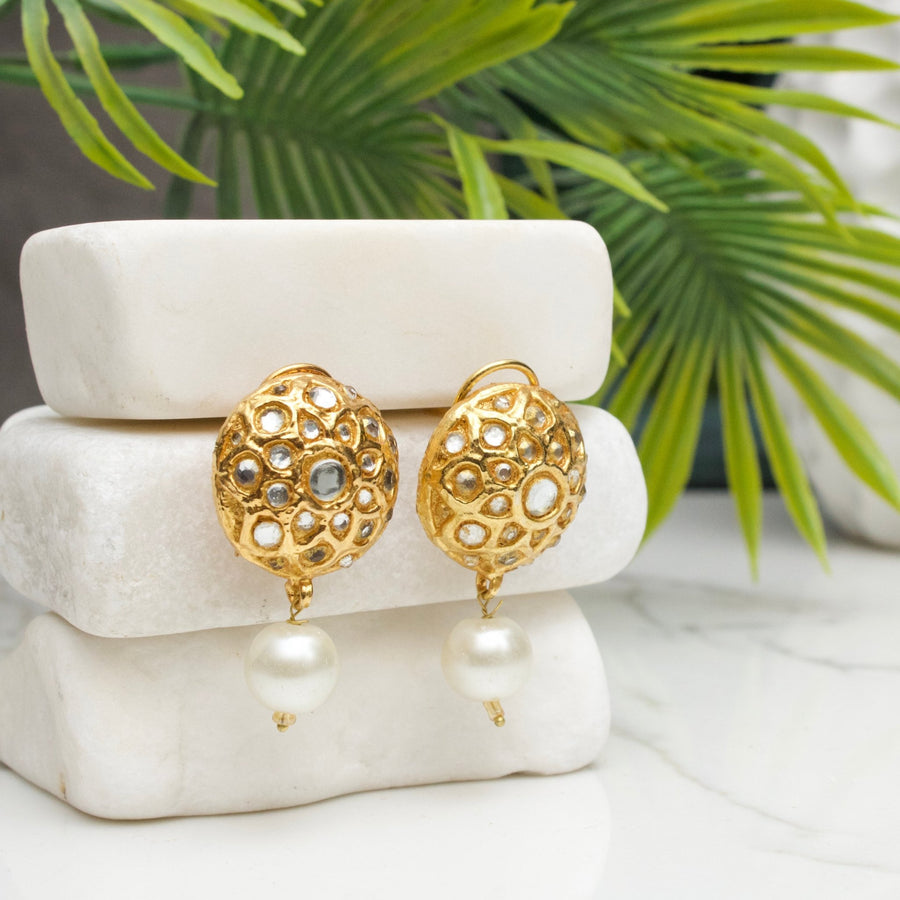 Pair of gold-tone drop earrings with intricate cut-out design and white pearl drop, displayed on white stone blocks with green foliage background.