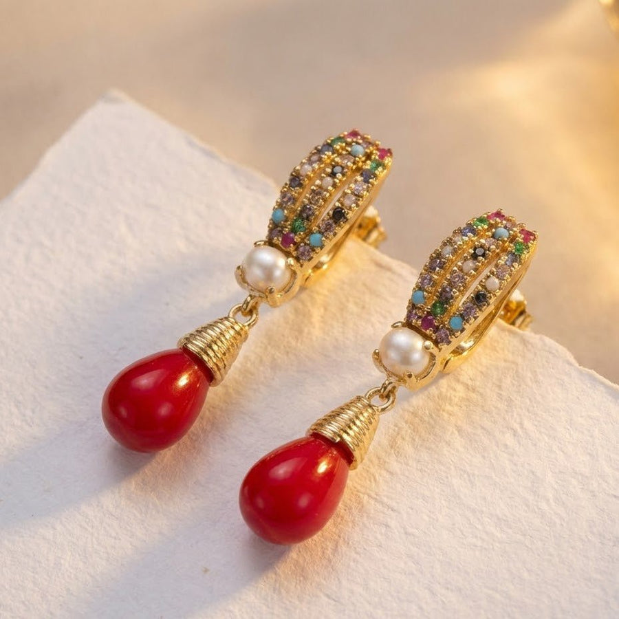 Close-up of gold-tone earrings featuring multicolor crystals, central pearl, and red teardrop pendant on a light background