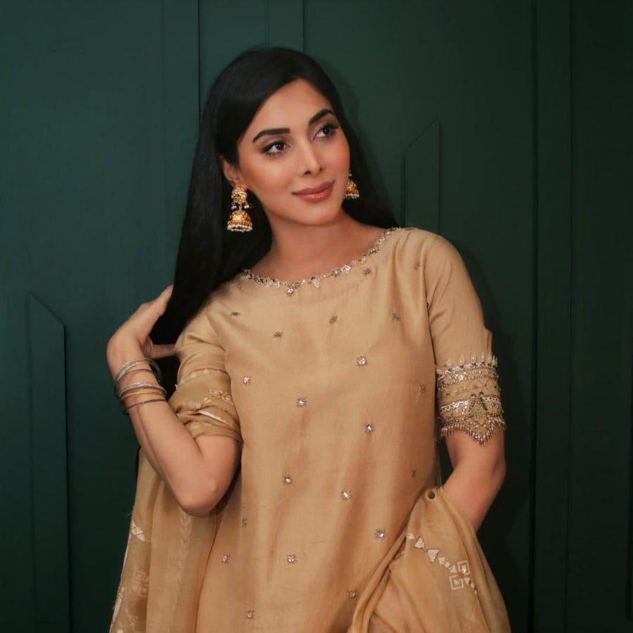 Woman wearing beige traditional outfit and elegant gold Jhumka earrings, posing with one hand touching her hair