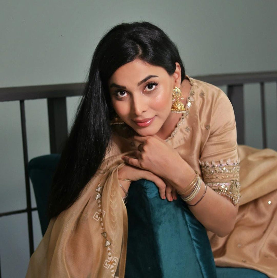 Woman in beige embroidered traditional dress leaning on a blue cushion, wearing statement gold Jhumka earrings with pearl details