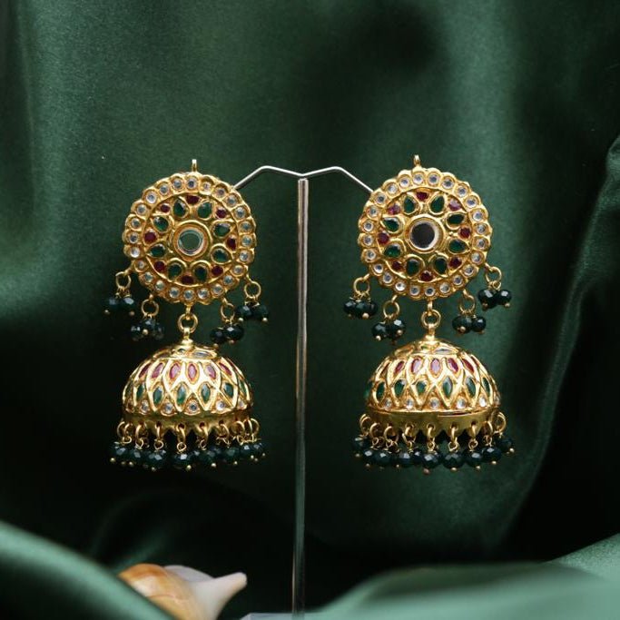 Pair of gold-plated Jhumka earrings decorated with multicolor precious stones and black beads displayed against a dark green fabric background