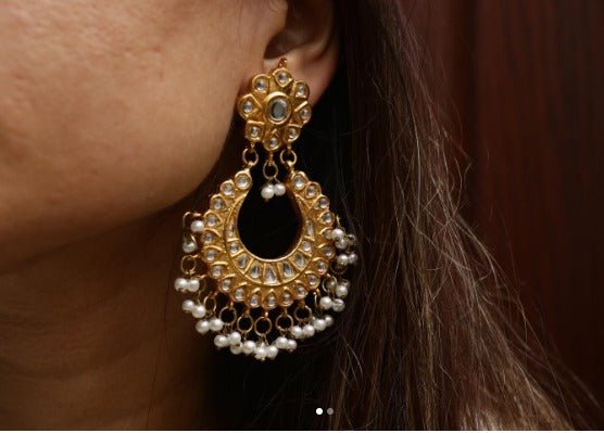 Close-up of a gold-plated Jhumka earring with clear stones and hanging white pearls worn on a woman's ear