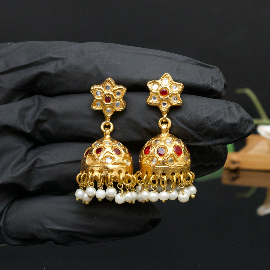 Hand holding gold-plated jhumka earrings with red gemstone accents, floral stud top, and pearl dangles against black background